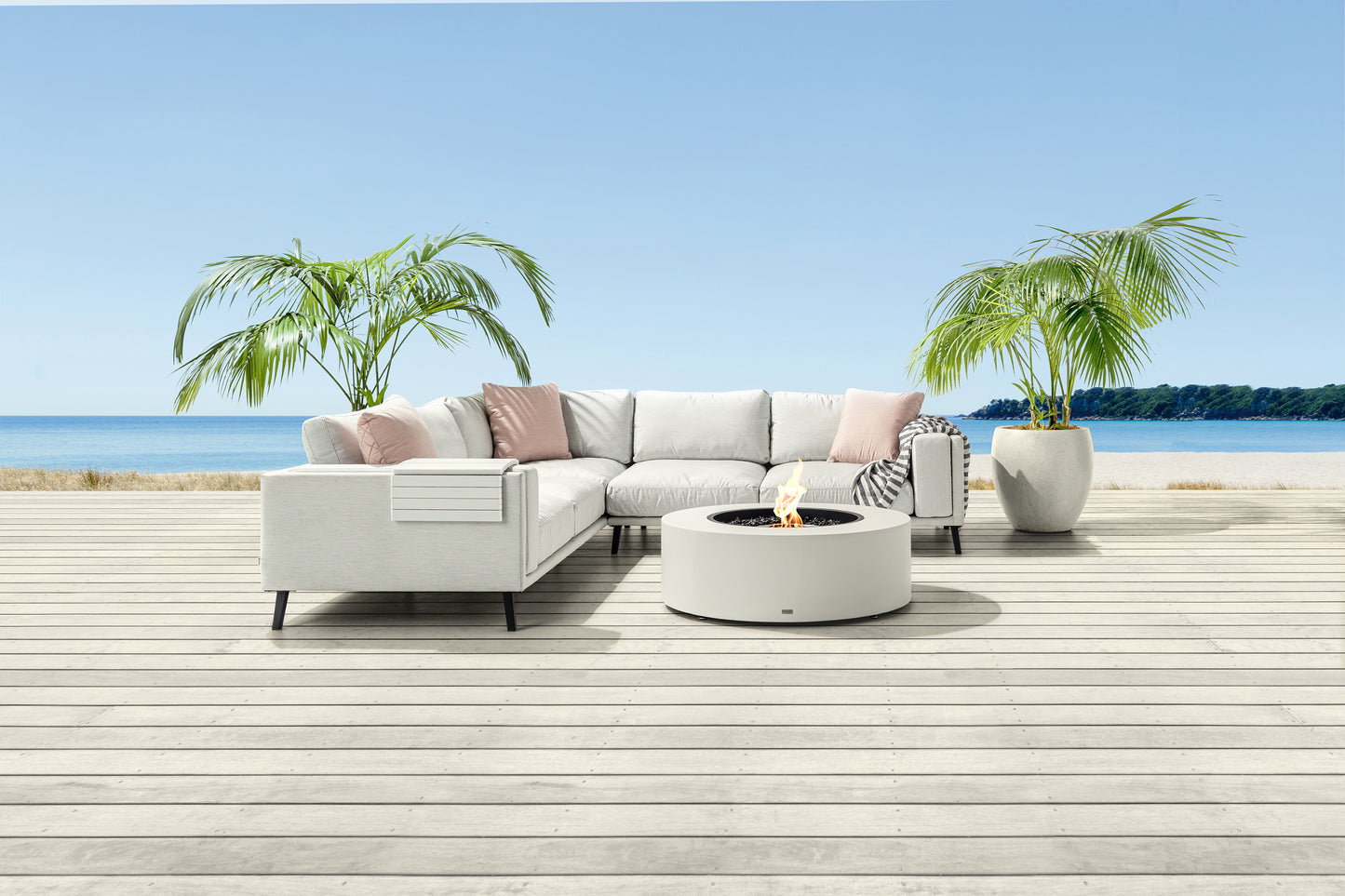 Outdoor patio set with sectional sofa, fire pit, and plants on a wooden deck by the water.