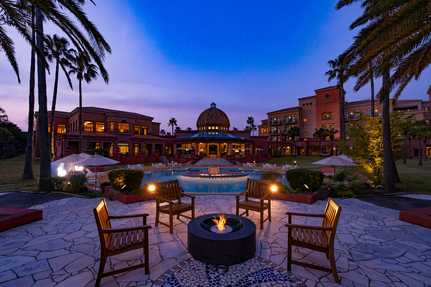 Evening scene at a resort with fire pit, chairs, and palm trees.