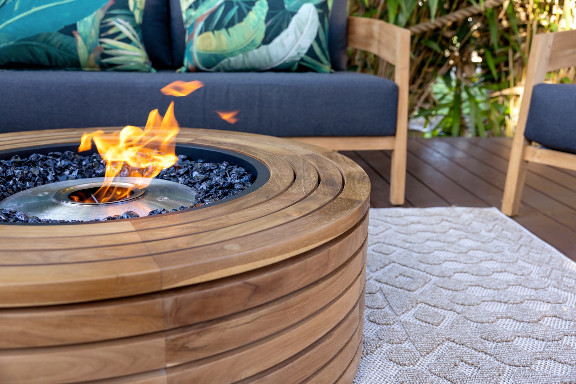 Wooden fire pit table with flames on a patio with outdoor furniture and greenery.