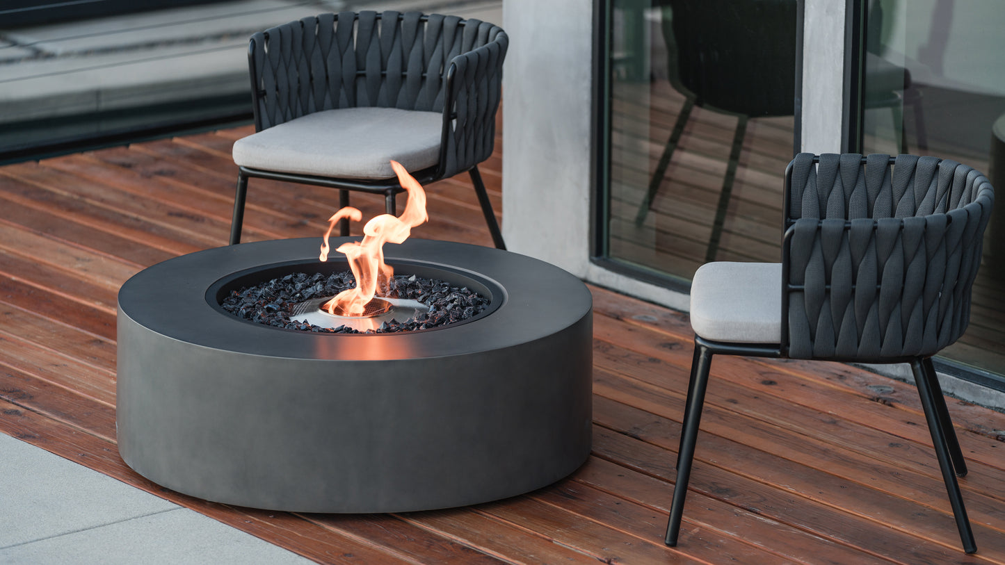 Round fire pit table with flames on a wooden deck, surrounded by two modern chairs.