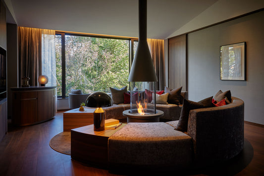 Modern living room with a fireplace, round ottoman, and large windows.