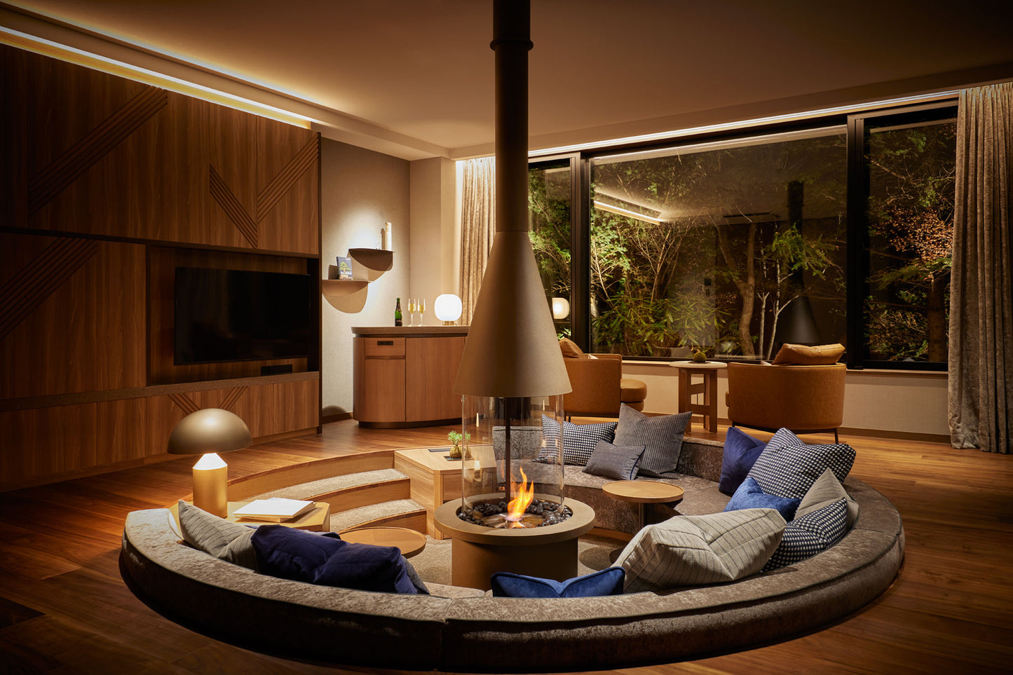 Modern living room with a circular sofa around a fire pit, large windows, and wooden furniture.