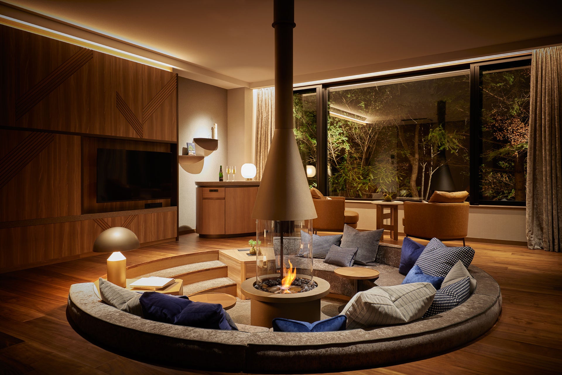 Modern living room with a circular sofa around a fire pit, large windows, and wooden furniture.