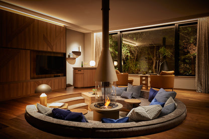 Modern living room with a circular sofa around a fire pit, large windows, and wooden furniture.