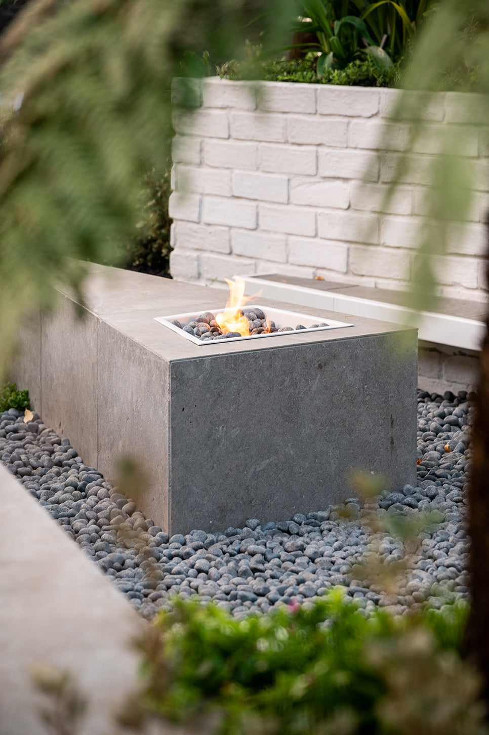 Concrete fire pit with flames surrounded by pebbles and greenery