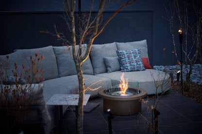 Outdoor seating area with a fire pit table and gray sofa against a dark wall.