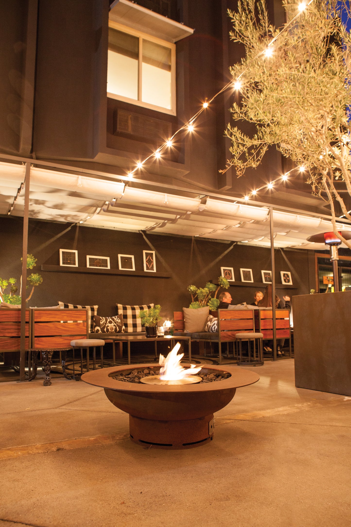 Outdoor patio with fire pit, string lights, and people sitting on a warm evening.