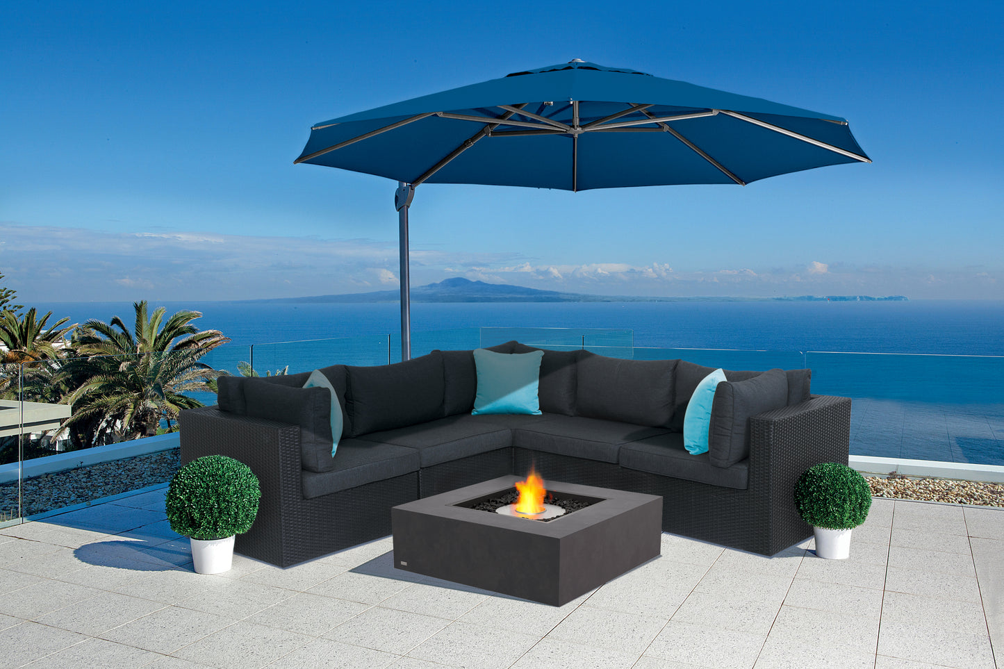 Outdoor patio set with black sectional sofa, fire pit table, and blue umbrella overlooking a scenic view.