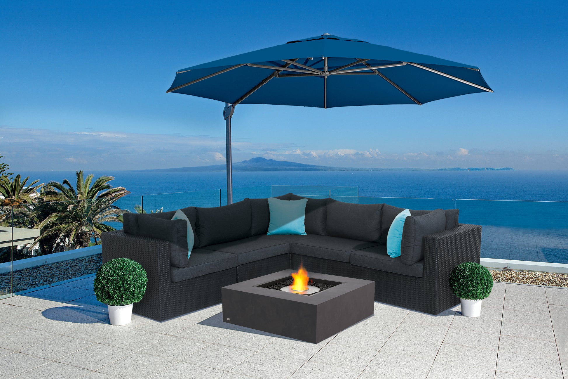 Outdoor patio set with black sectional sofa, fire pit table, and blue umbrella overlooking a scenic view.