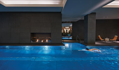 Person swimming in an indoor pool with a modern design featuring a fireplace.