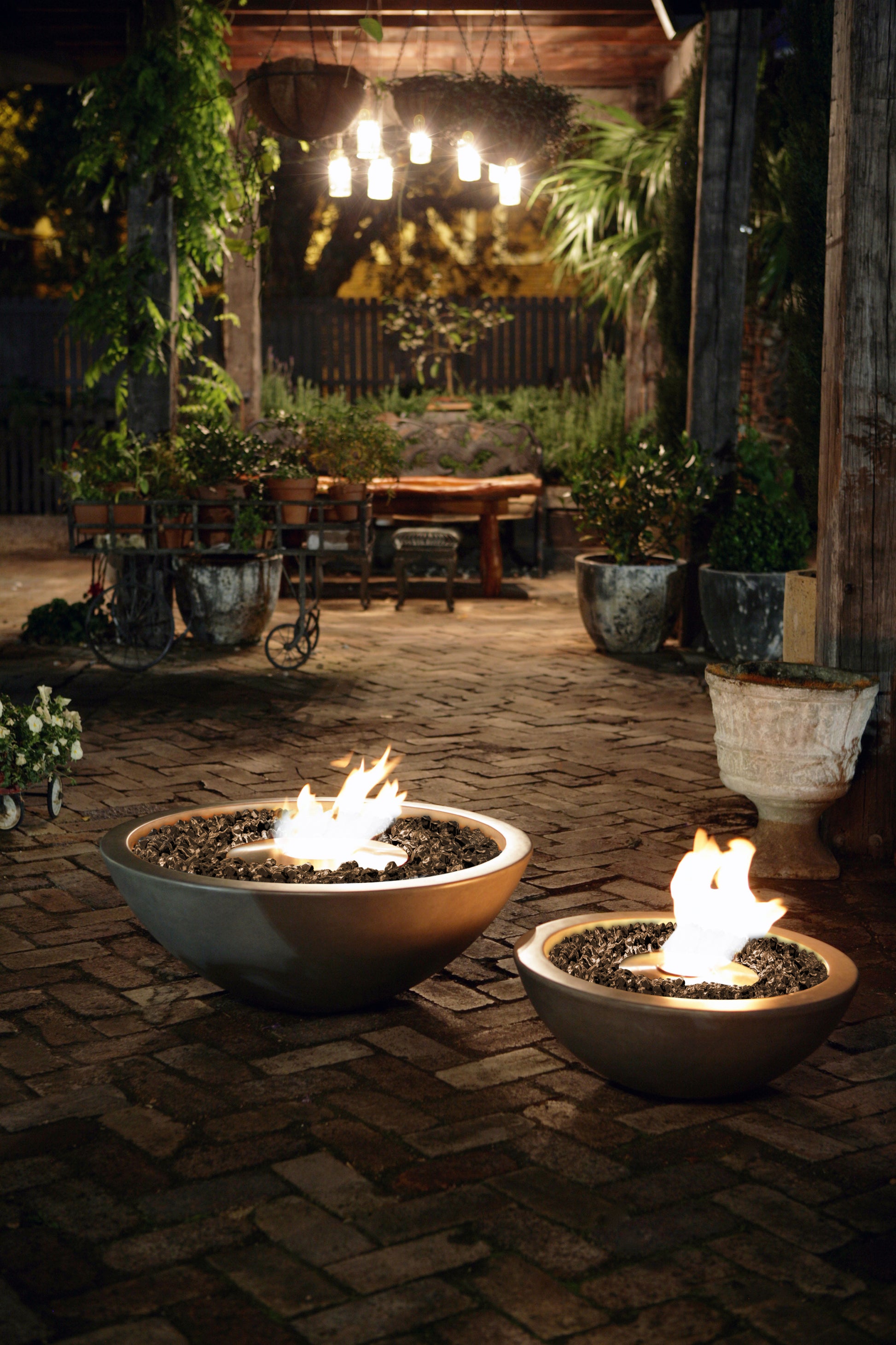 Two fire pits with flames on a stone patio at night, surrounded by potted plants and a wooden table.