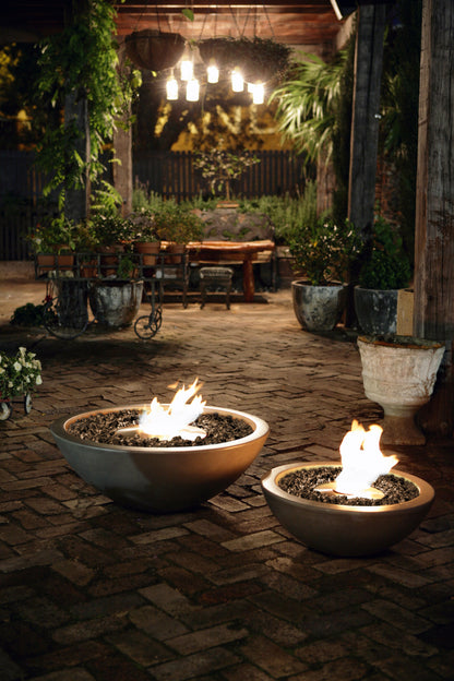 Two fire pits with flames on a stone patio at night, surrounded by potted plants and a wooden table.