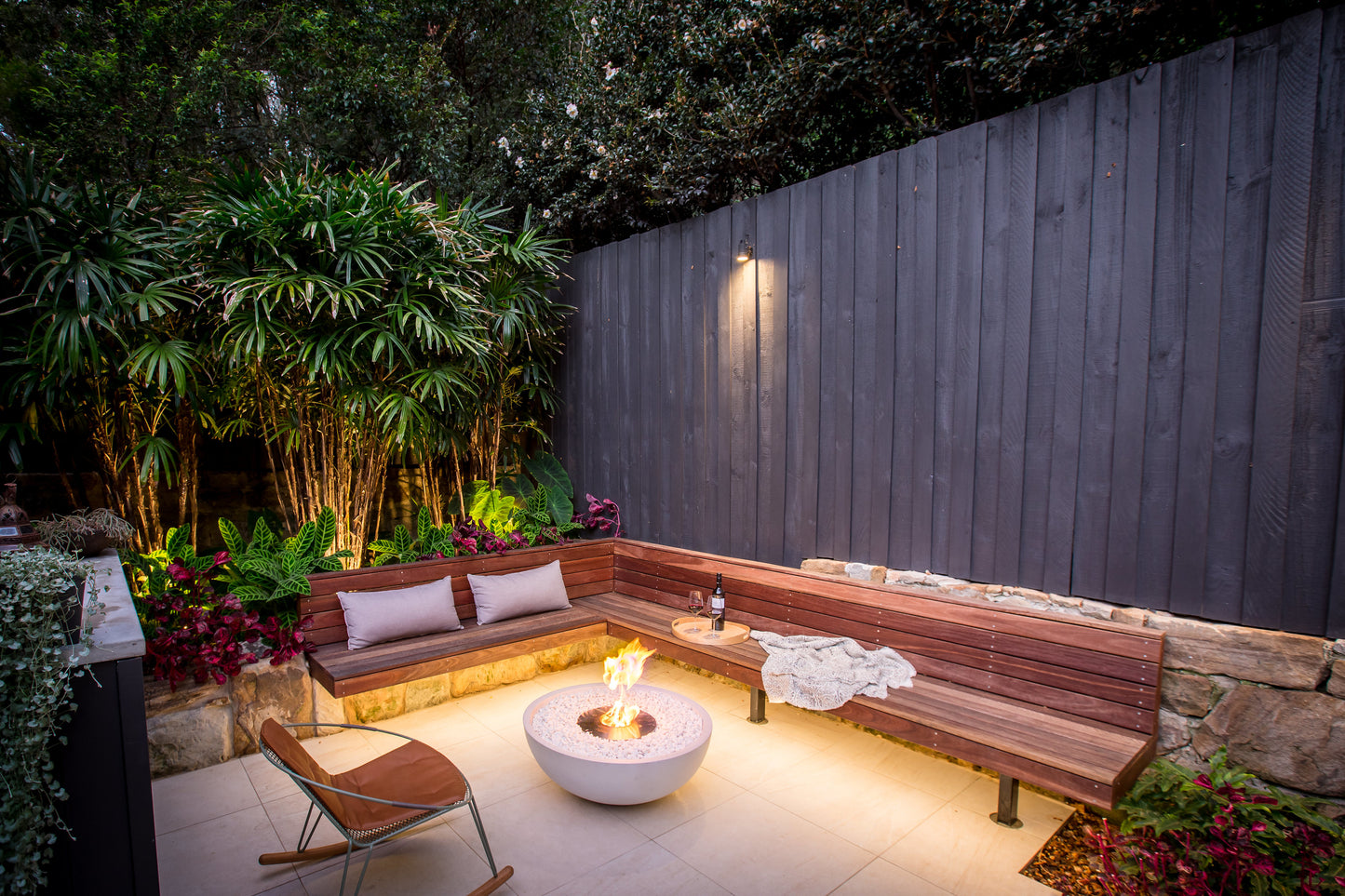 Outdoor seating area with a fire pit, wooden bench, and chairs in a garden setting.