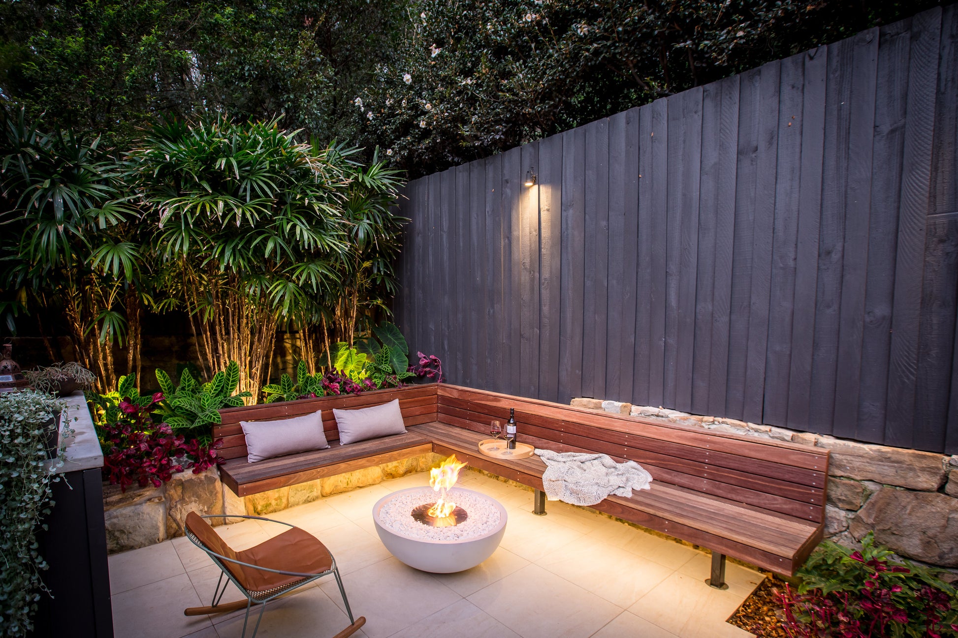Outdoor seating area with a fire pit, wooden bench, and chairs in a garden setting.