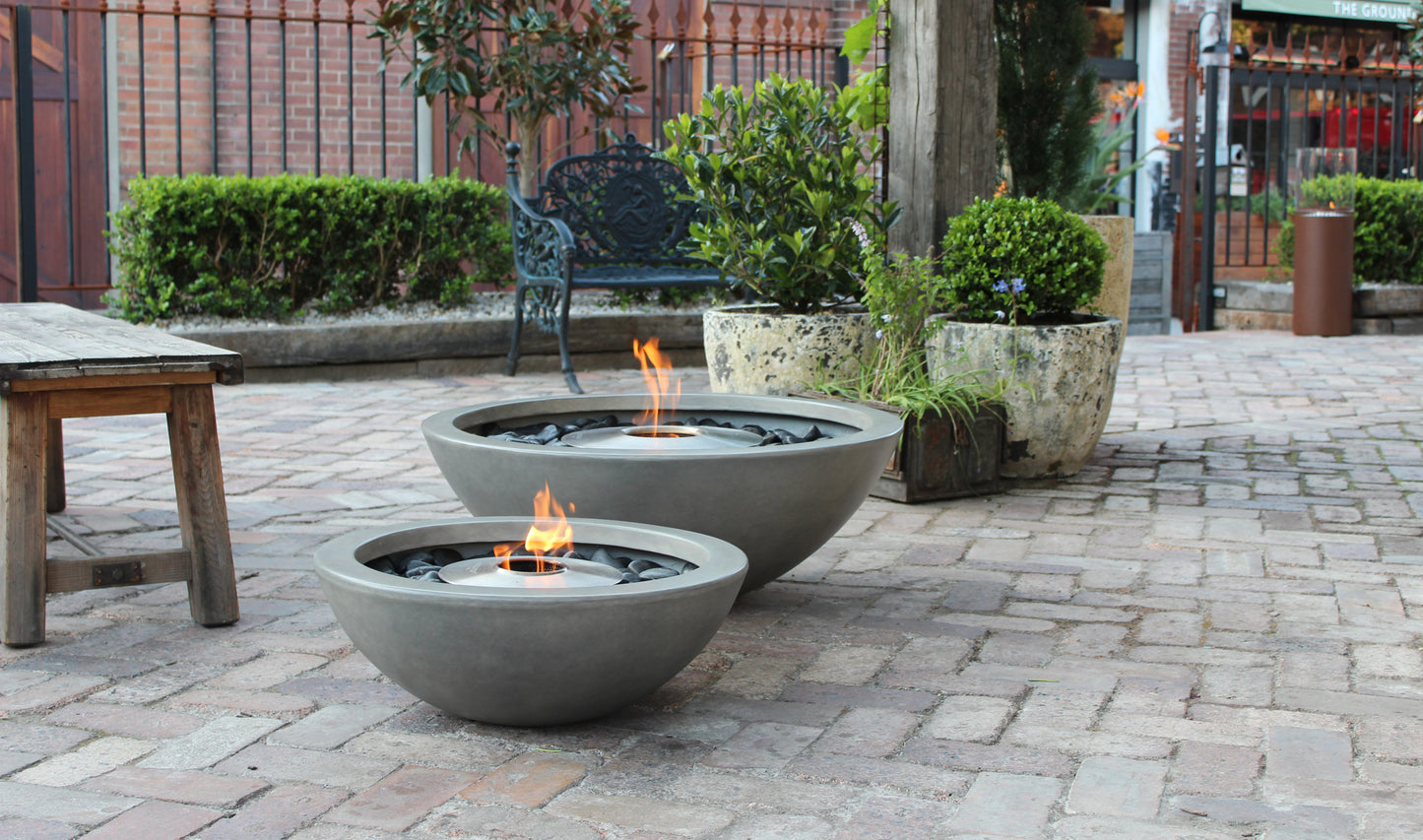 Two fire pits on a patio with flames lit, surrounded by potted plants and a bench.