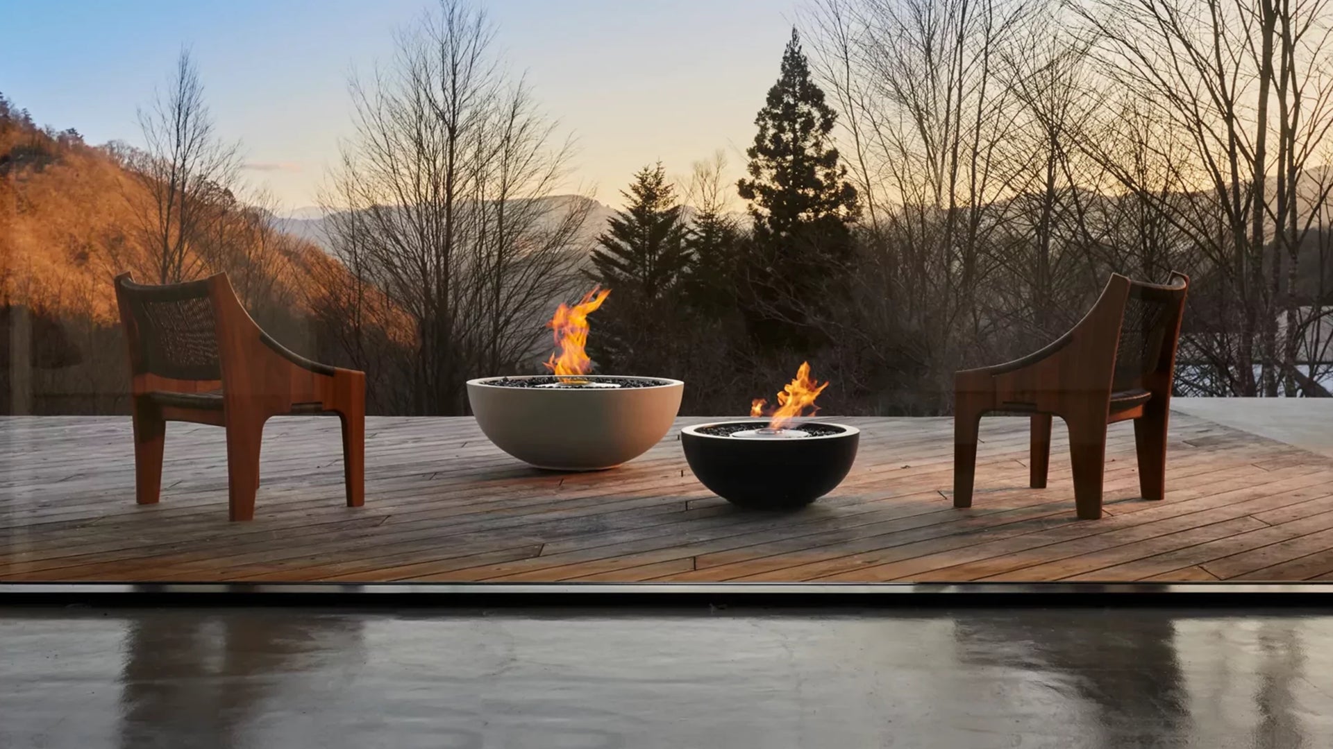 Two wooden chairs around two fire pits on a wooden deck with a scenic background.