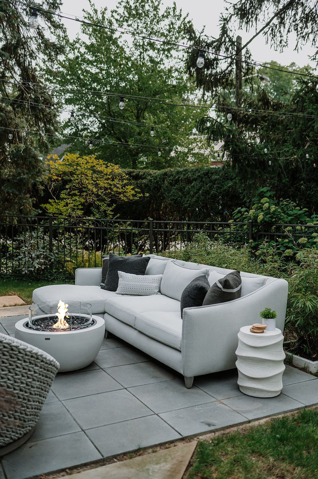 Outdoor patio with a white sectional sofa, fire pit, and small table in a garden setting.