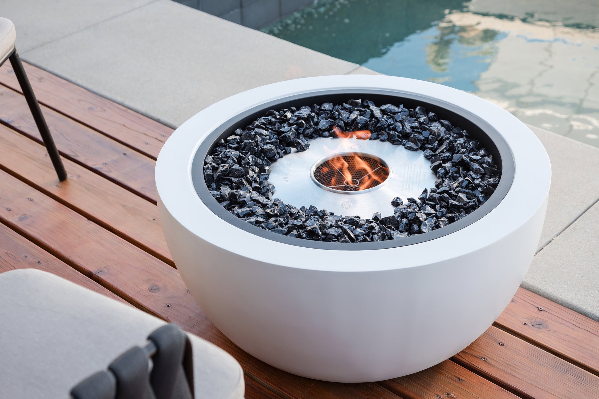 White fire pit table with flames on a wooden deck by a pool