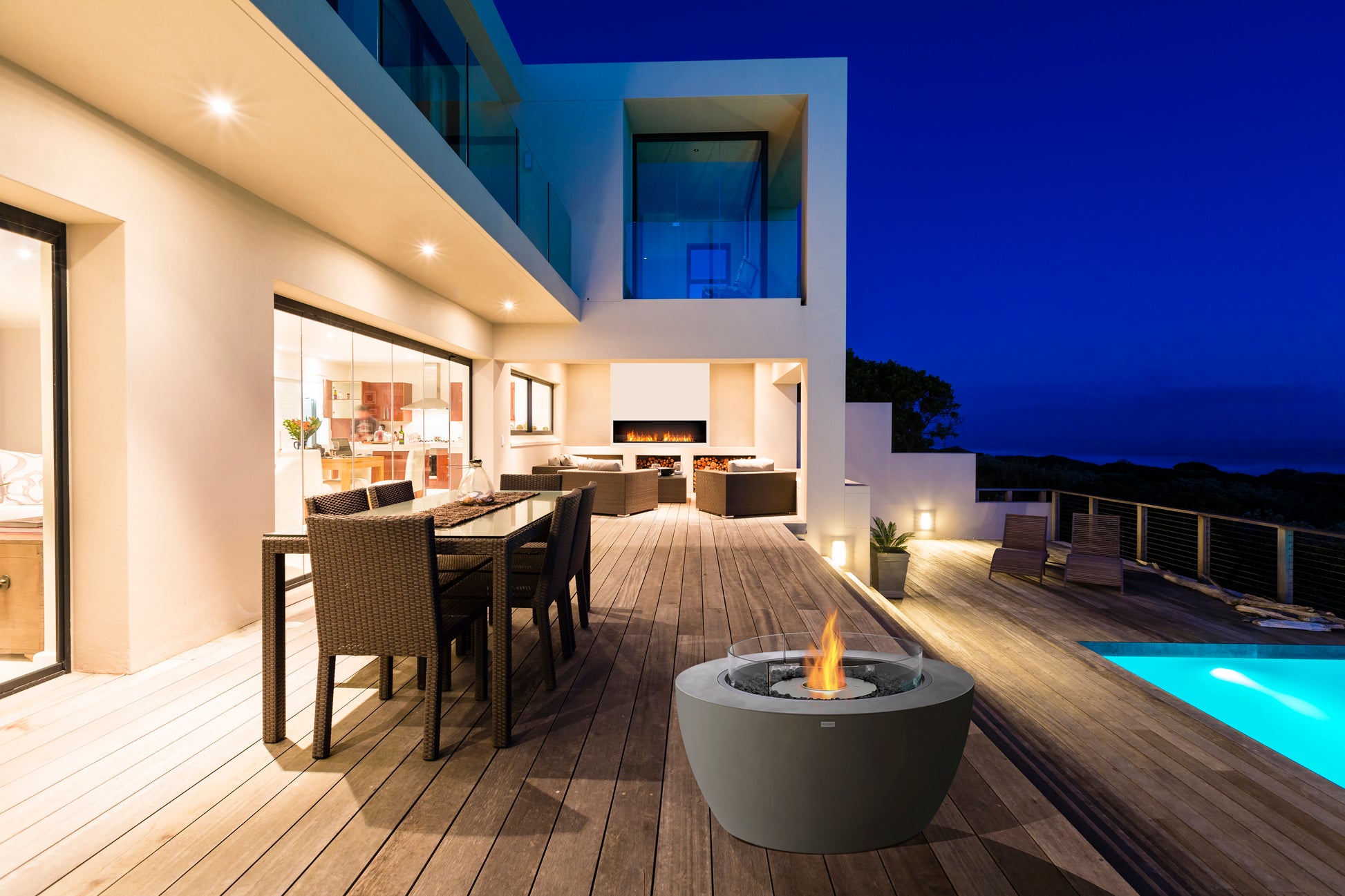 Modern outdoor patio with dining area, fire pit, and pool at night.