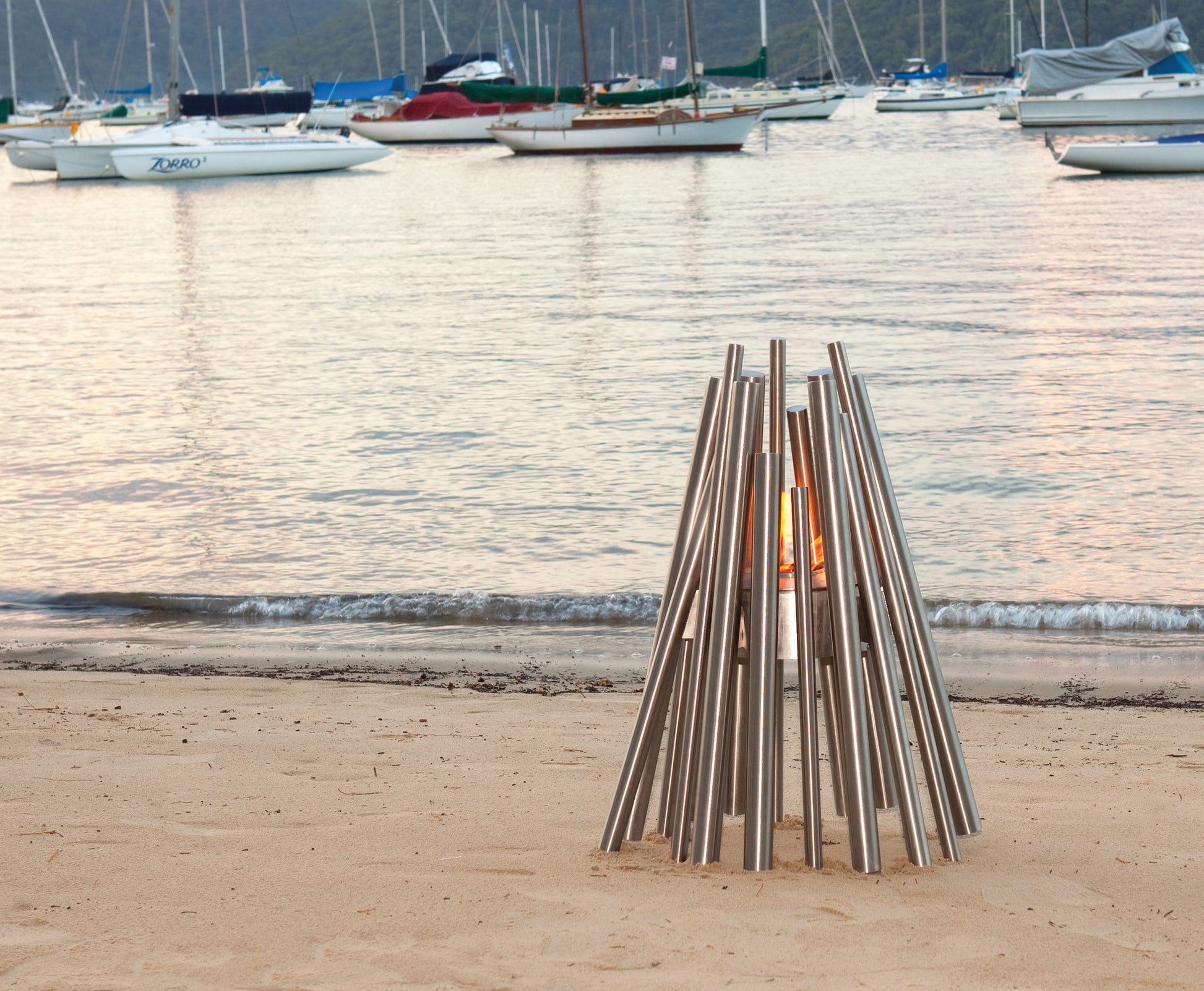 Modern outdoor fire pit on a sandy beach with boats in the background