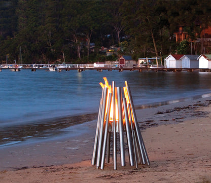 Metal fire pit on a beach with a scenic background of water and trees.