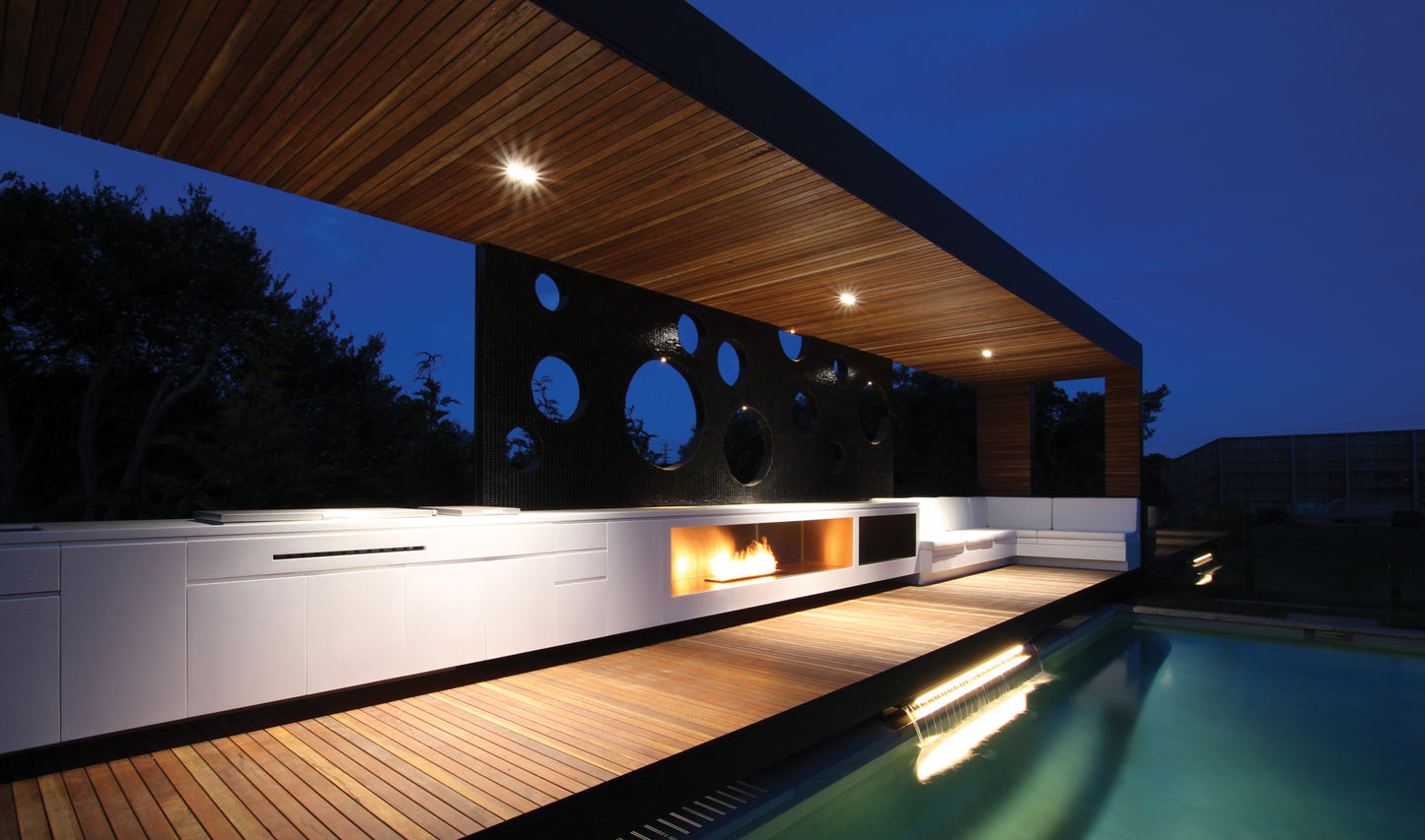 Modern outdoor living space with a pool, fire pit, and wooden deck at night.