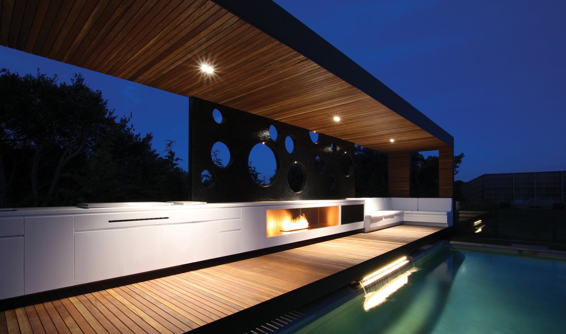 Modern outdoor living space with a pool, fire pit, and wooden deck at night.